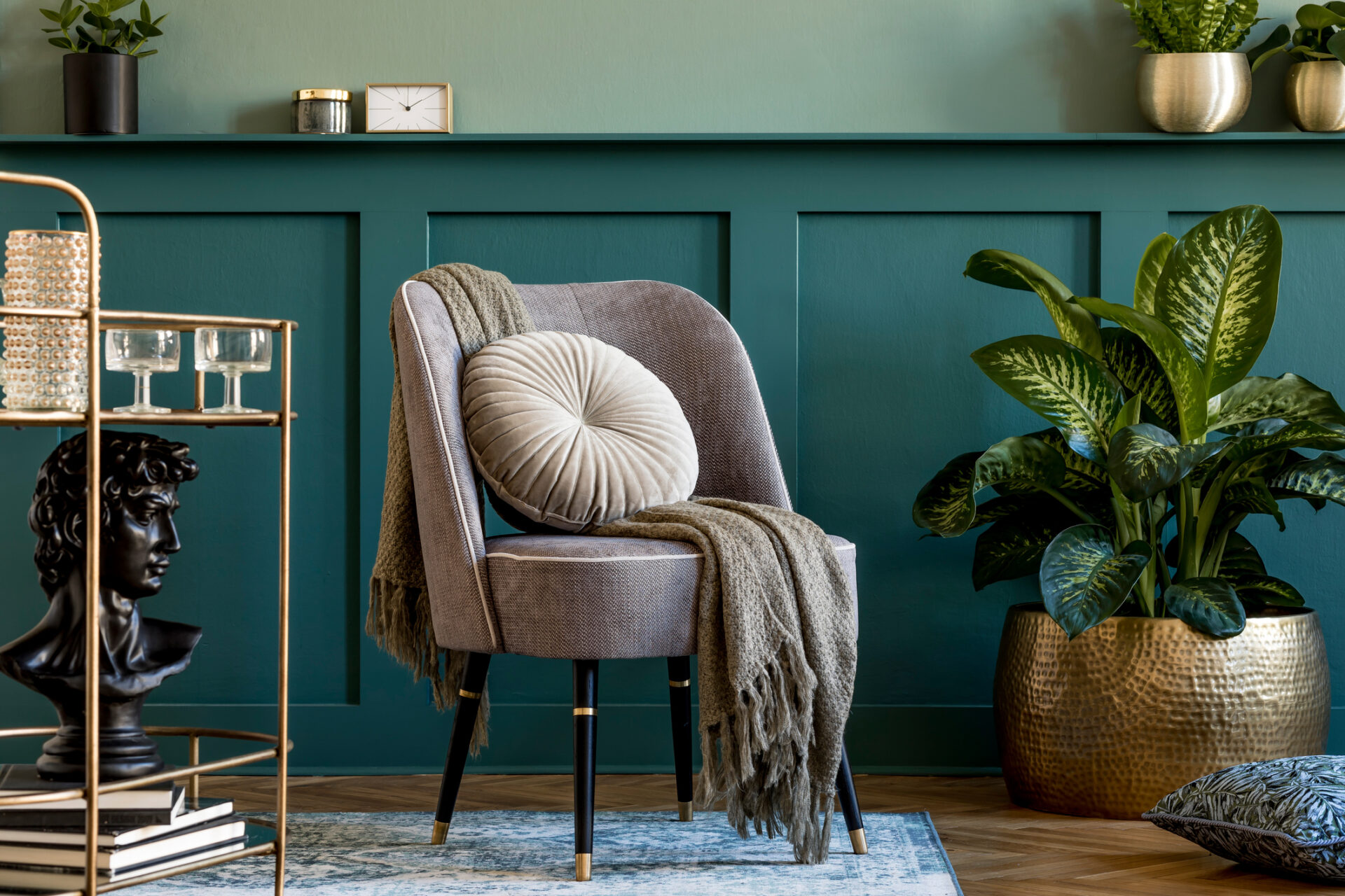 Stylish and modern composition of living room with design gray armchair, gold liquor cabinet, plants and elegant personal accessories. Gray wall panelling with shelf. Modern home decor. Template.